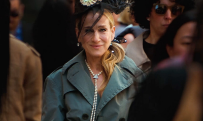 Sarah Jessica Parker wears gray trench coat, black veiled hat, and Ben-Amun pearl necklace surrounded by a crowd of people walking