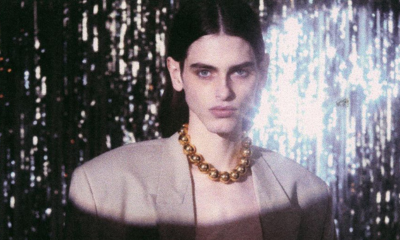 Individual wears gold 8th & 38th Collection necklace from Ben-Amun with an oversized blazer and middle-part hair against a silver streamer background